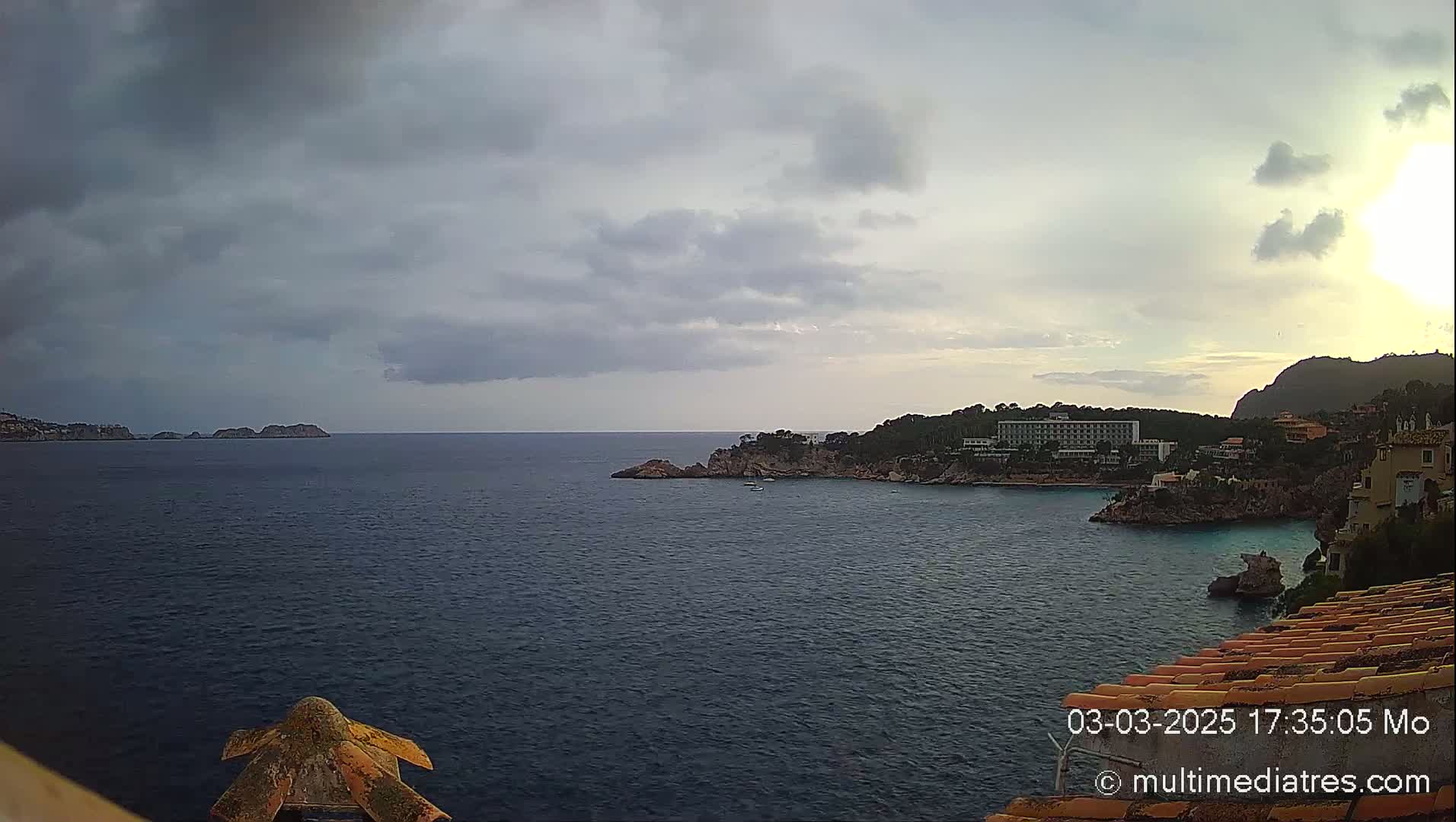 A partly cloudy coastal scene shows a dark blue sea with rocky shorelines and buildings, including a large hotel, under a mostly gray sky.