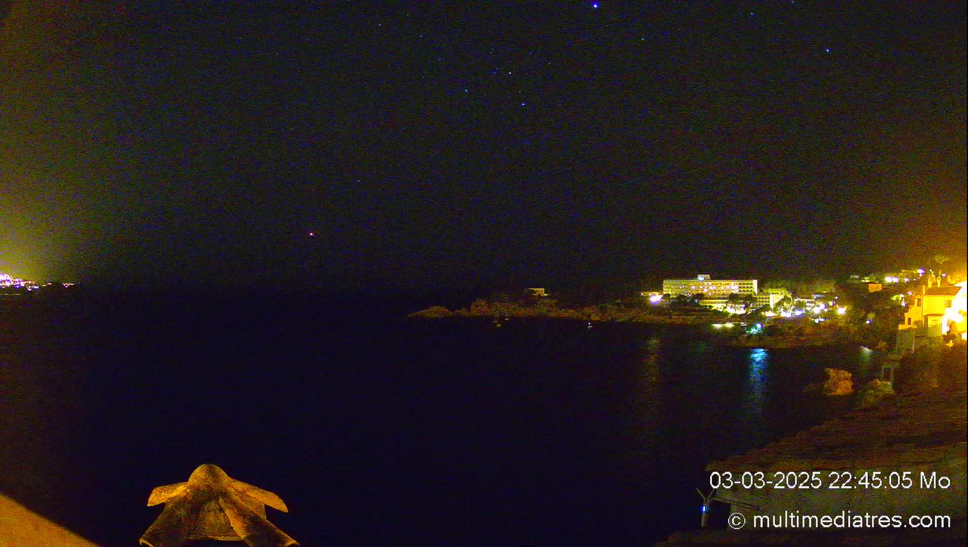 A nighttime view of a calm body of water with a brightly lit hotel and buildings on the opposite shore under a clear, starlit sky.