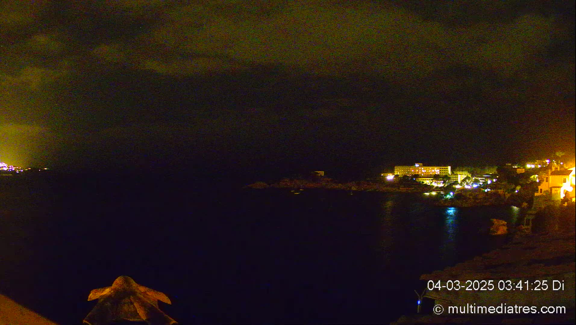 A nighttime coastal scene shows a dark body of water reflecting the lights of buildings on the shore under a cloudy sky.