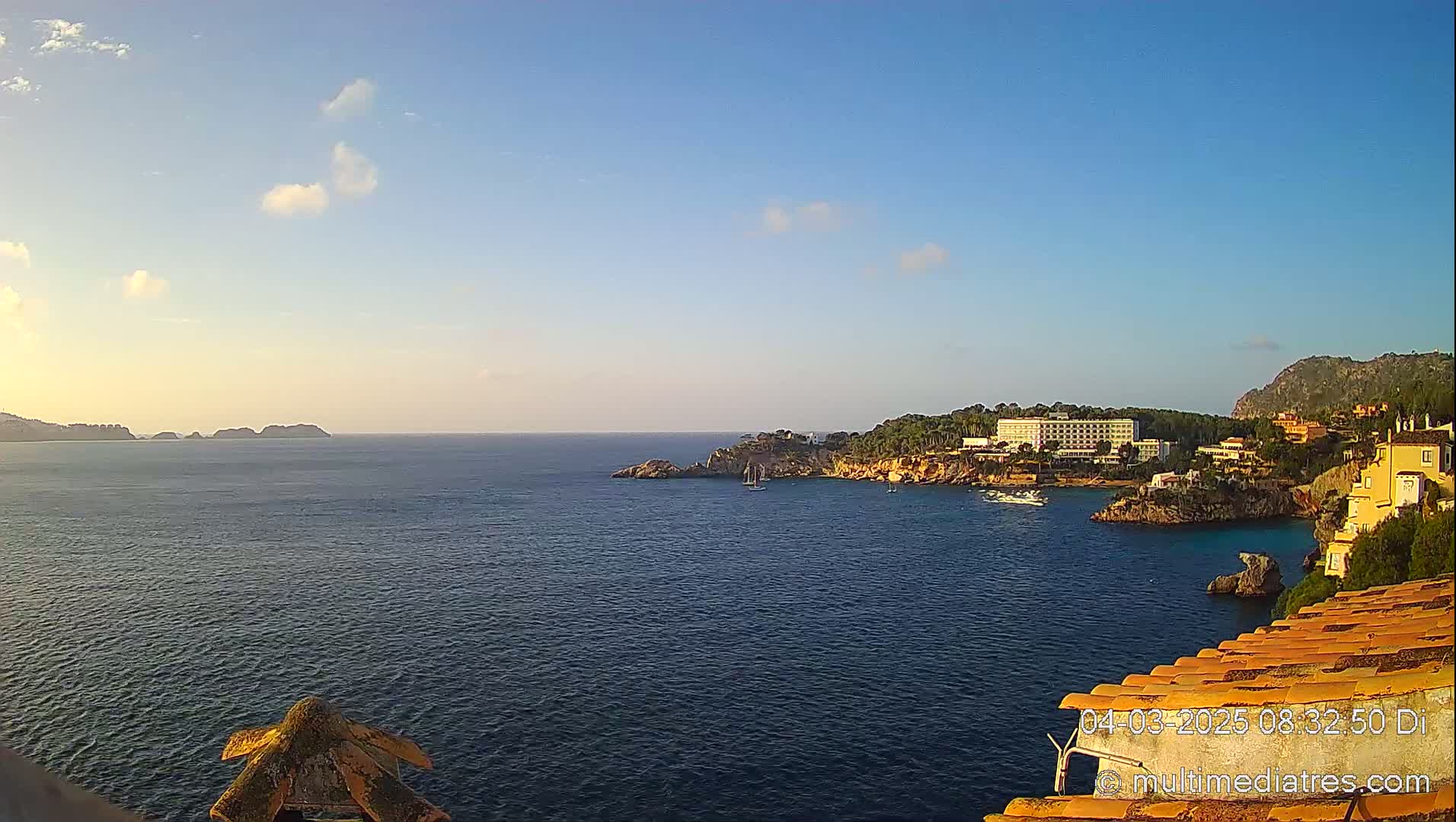 A calm, clear day reveals a coastal scene with a hotel and buildings nestled on a rocky shoreline, overlooking a tranquil bay with a few boats, and distant islands under a pale blue sky.