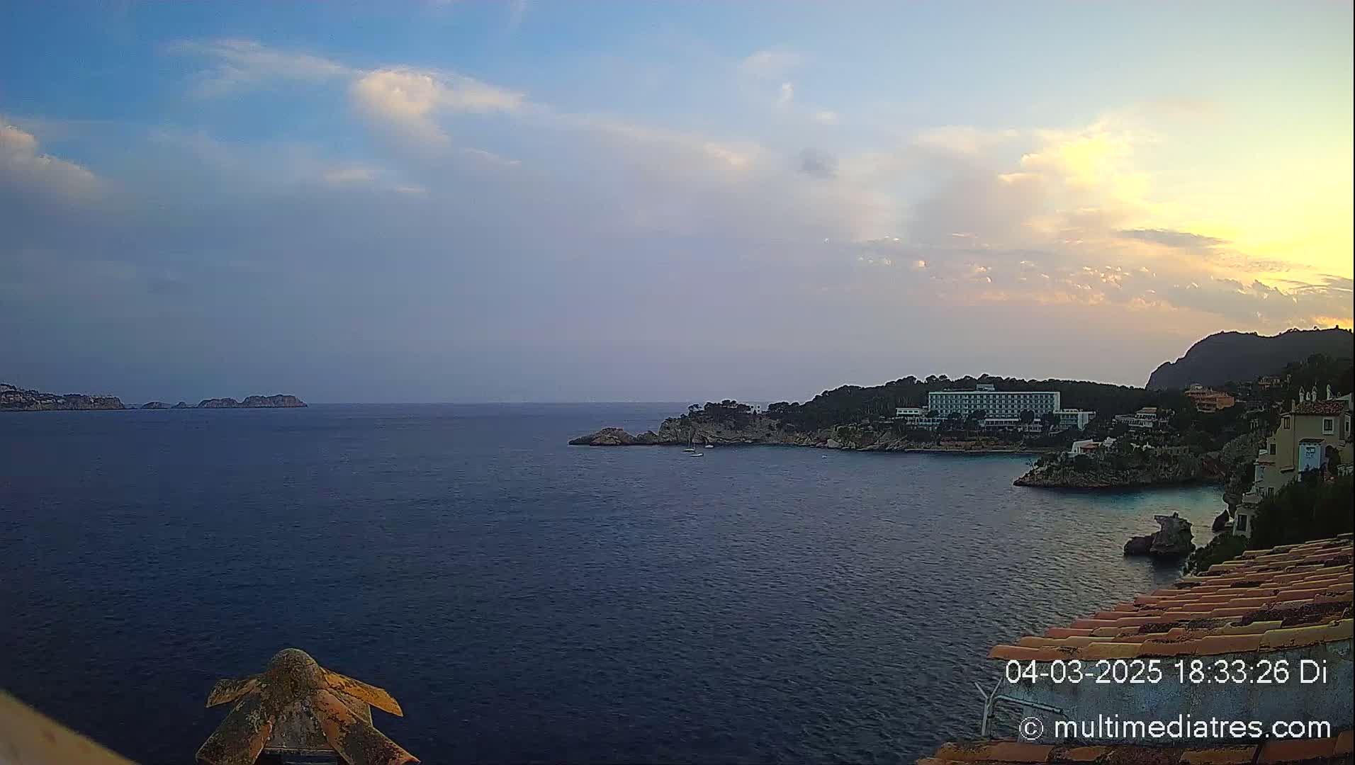 A tranquil coastal scene at sunset, featuring a calm sea, rocky shores, and a large hotel nestled amongst the trees.