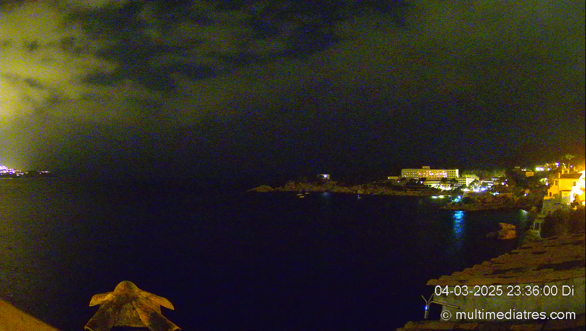 A nighttime coastal scene shows a dark body of water with a lit-up shoreline featuring buildings and some boats, under a mostly cloudy sky.