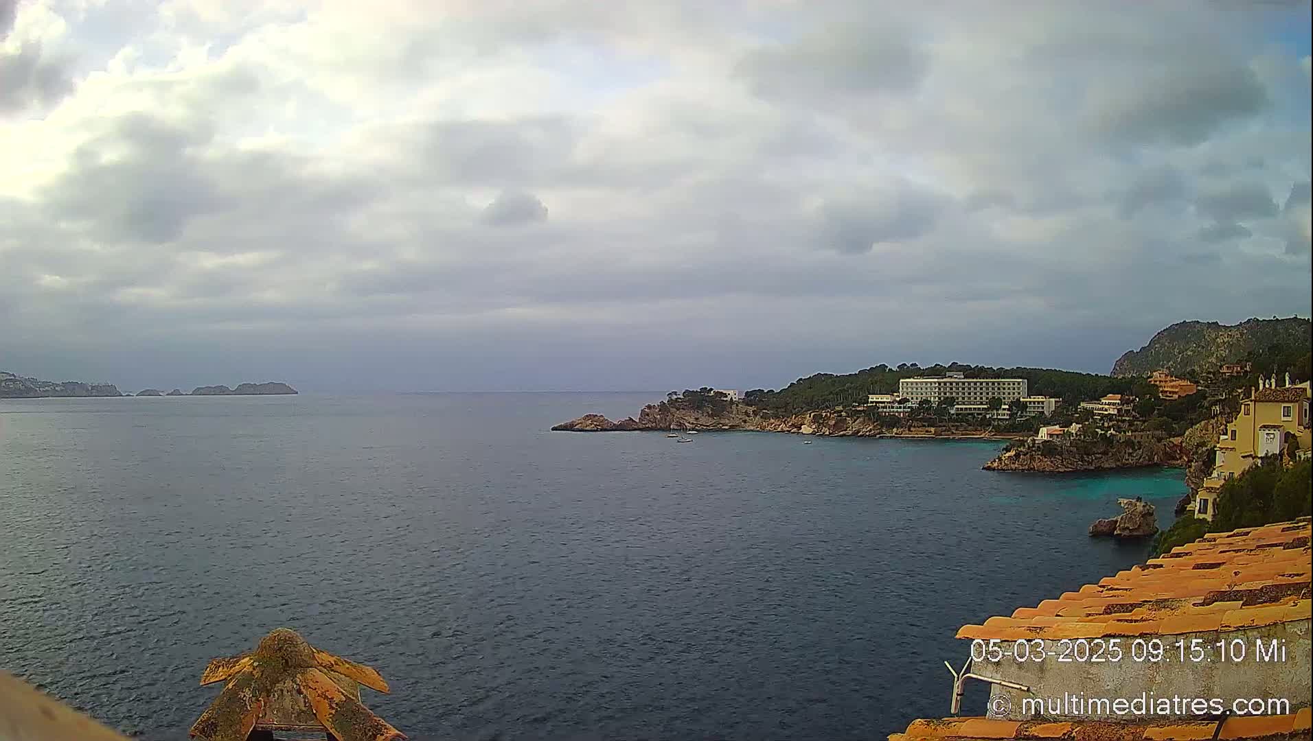 A coastal scene shows a calm sea under a cloudy sky, with a hotel and other buildings situated on a rocky shoreline.