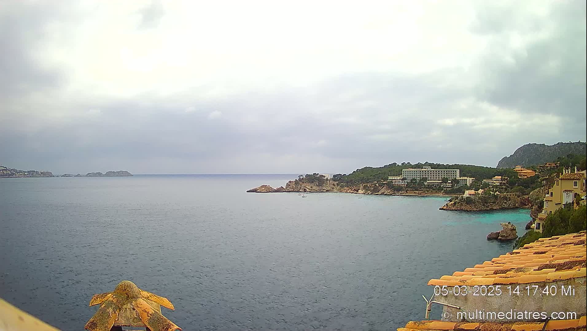 A coastal view on an overcast day shows a calm sea with rocky outcrops, a hotel, and buildings on a hillside.