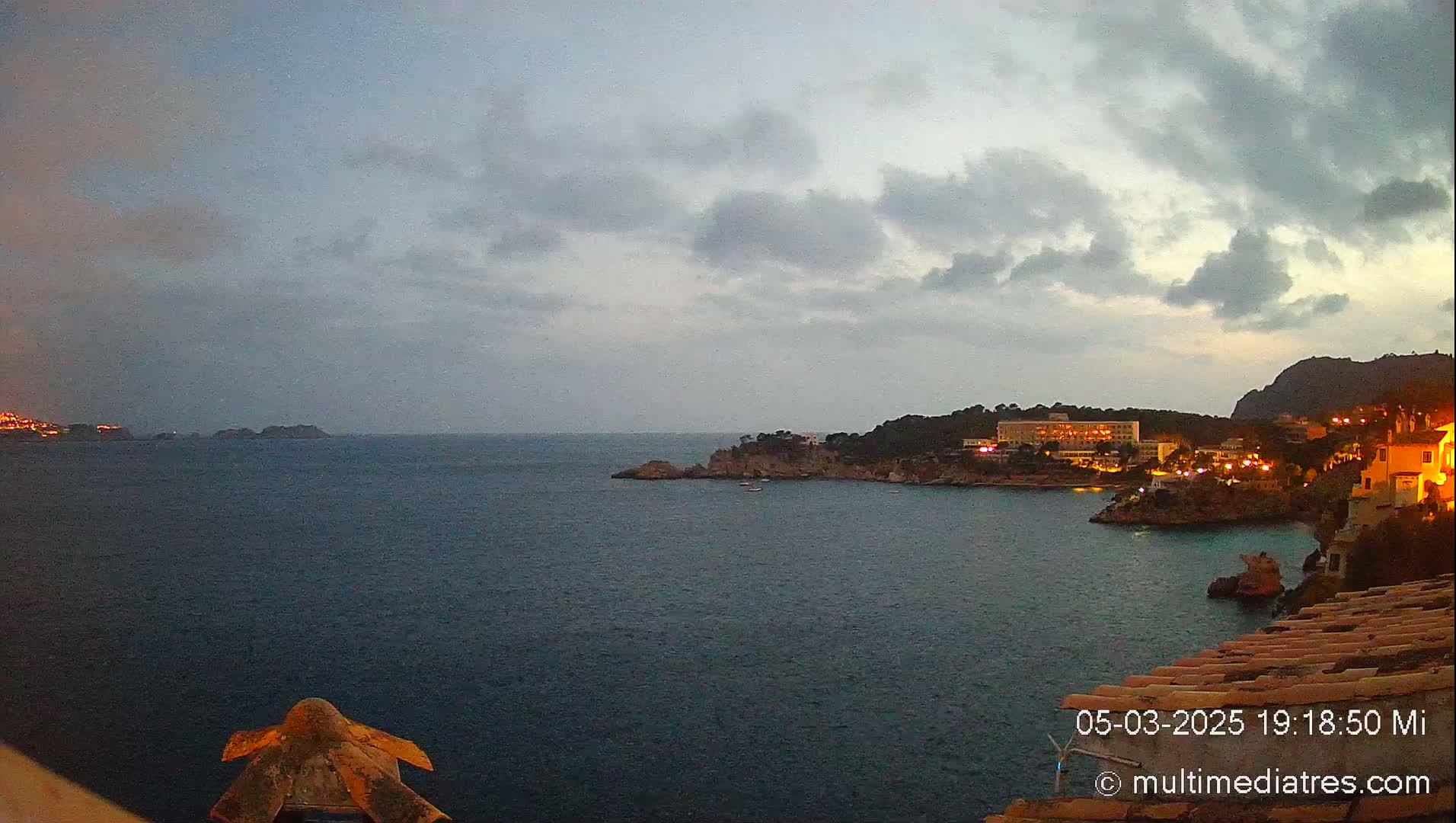 A twilight view of a calm, dark-blue sea with illuminated buildings and rocky coastlines under a mostly cloudy sky.