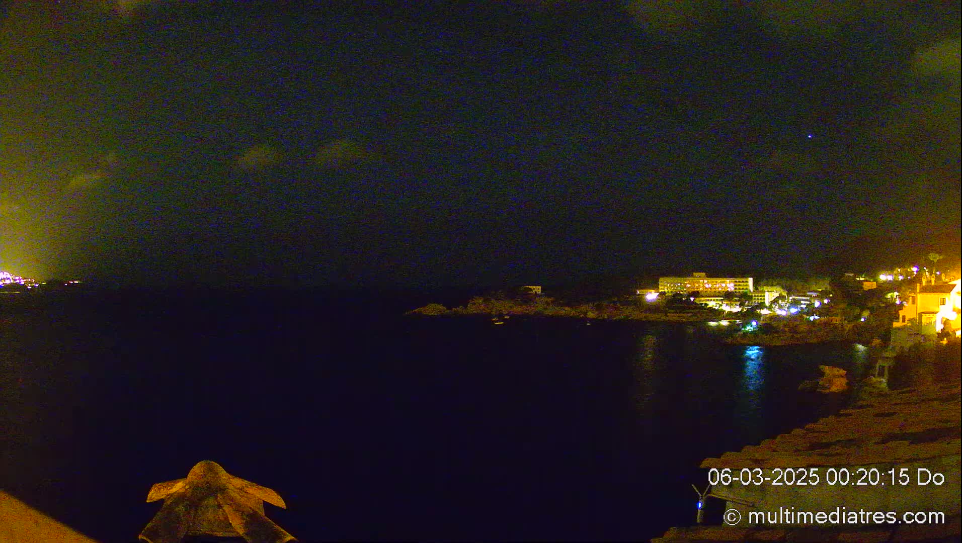 A nighttime view of a dark, calm body of water with illuminated buildings on the far shore under a mostly clear sky.