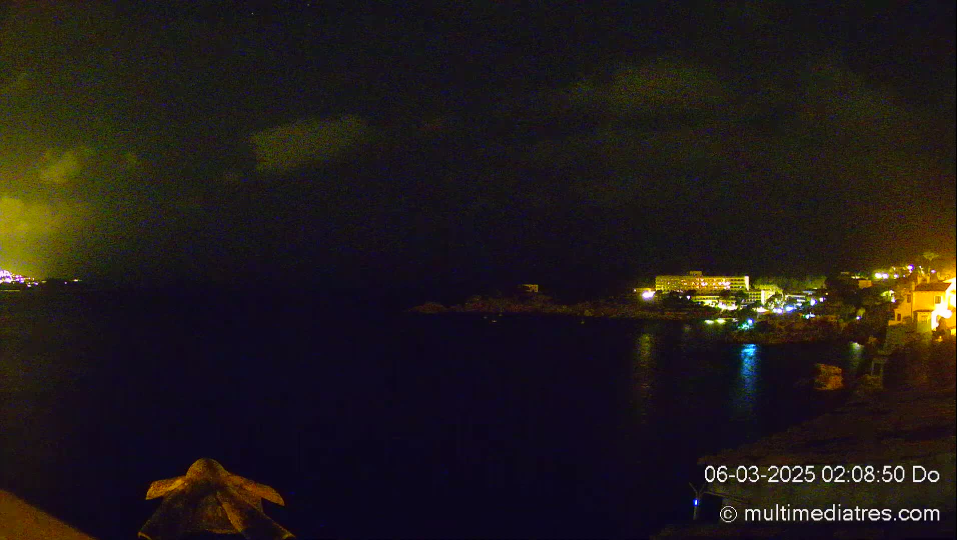 A nighttime view of a dark, calm body of water with illuminated buildings and a partly cloudy sky visible in the distance.