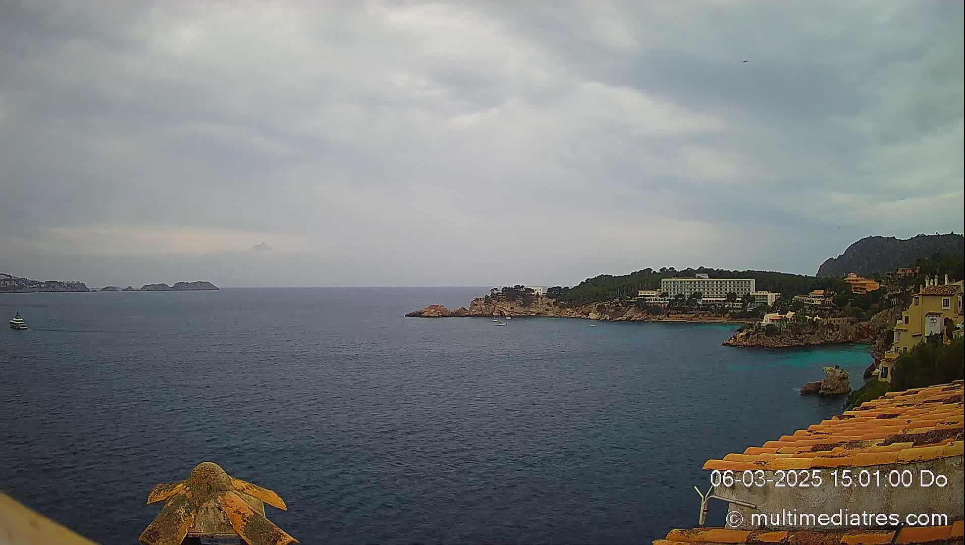 A tranquil coastal scene under a cloudy sky shows a body of water with a boat in the distance, rocky shorelines, and buildings nestled amongst greenery on the hillsides.