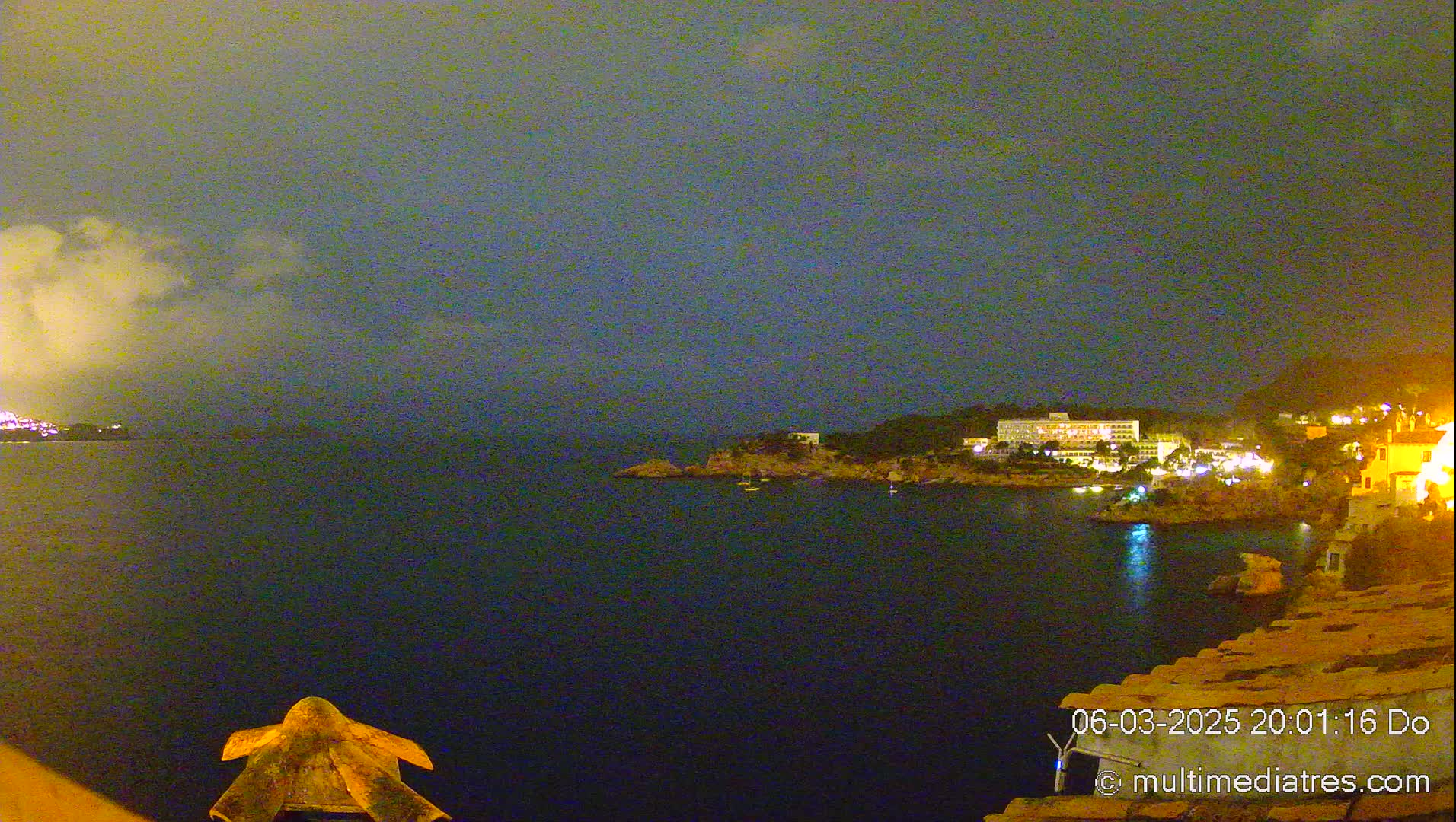 A nighttime coastal scene shows a dark body of water with a faintly lit shoreline featuring buildings, under a cloudy, dark sky.