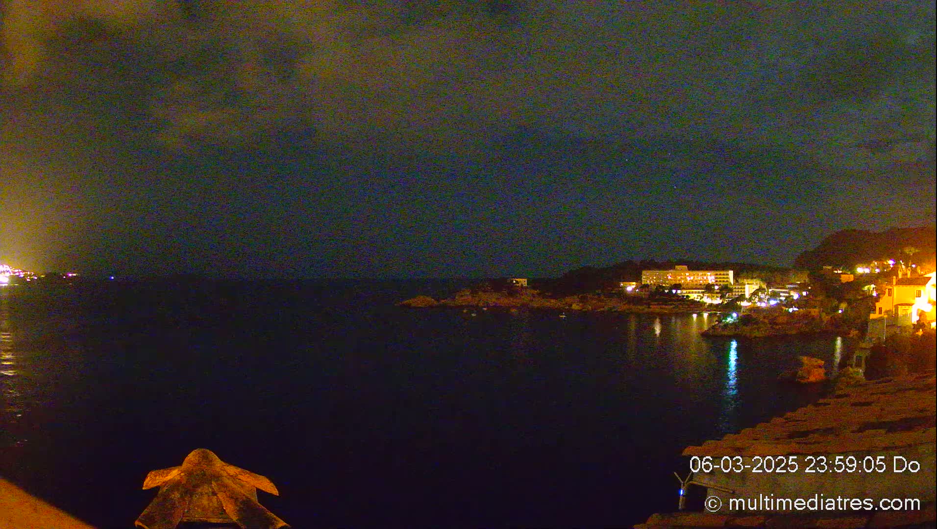 A nighttime view of a dark, calm body of water with illuminated buildings and land on the opposite shore under a cloudy sky.