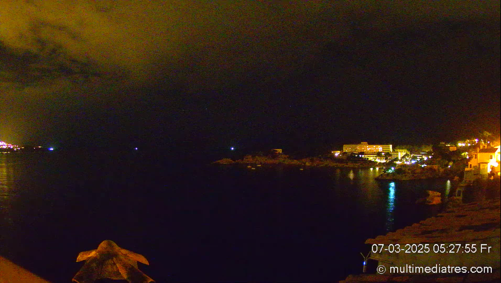 A nighttime view of a calm body of water with illuminated buildings and a few small lights on the opposite shore under a dark, cloudy sky.