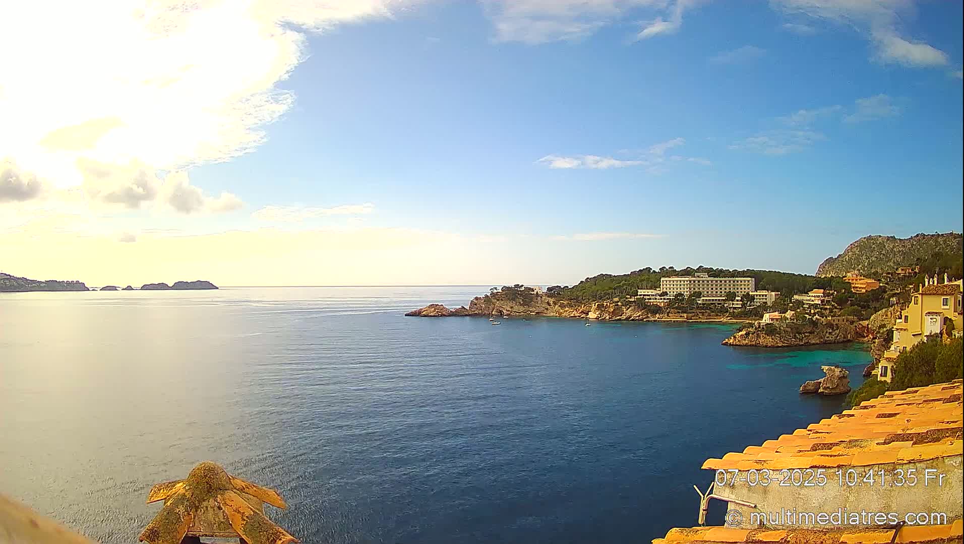 A partly cloudy, sunny day overlooks a calm ocean bay with a hotel and buildings on a rocky coastline and distant islands.