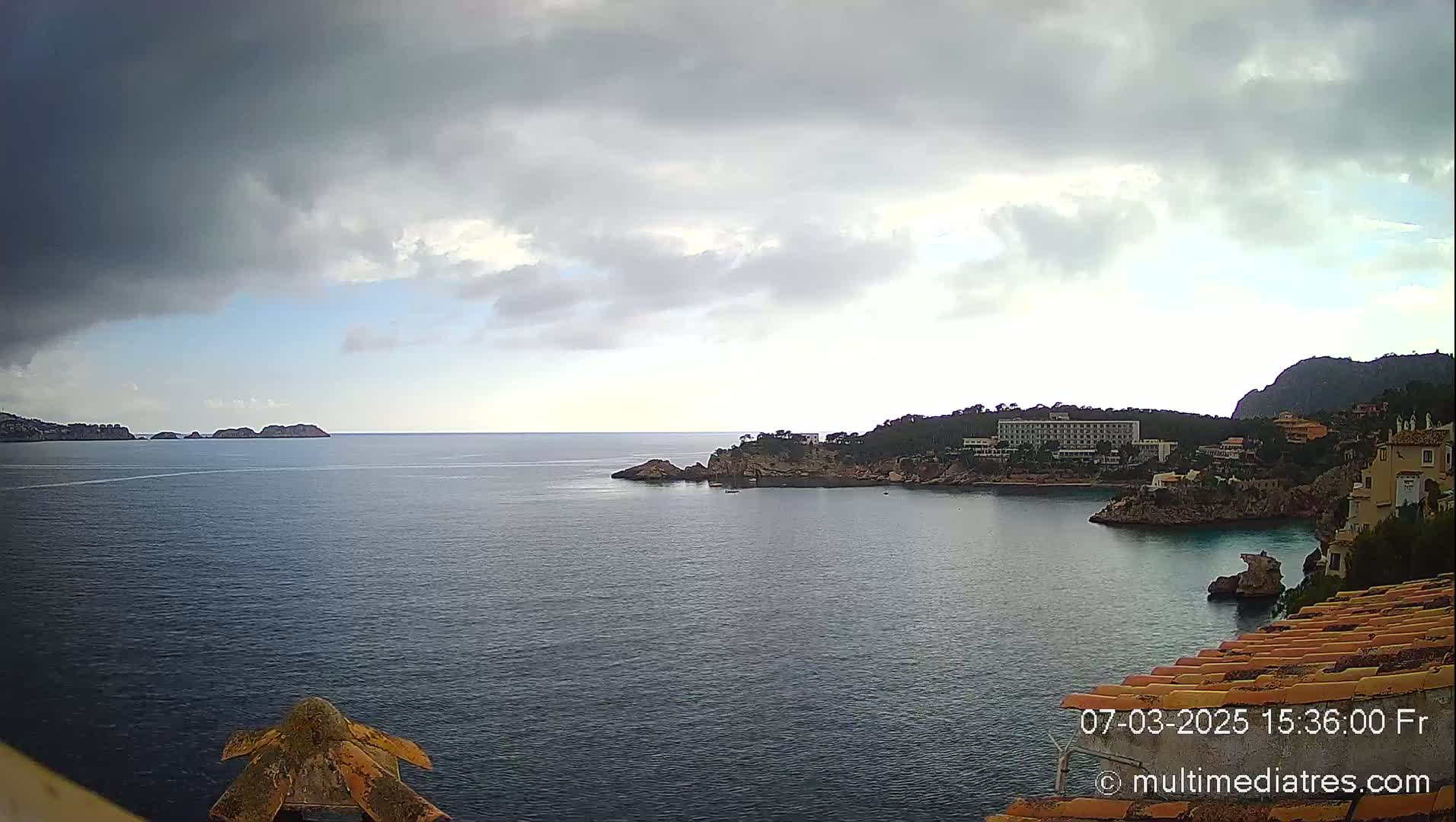 A coastal scene under a mostly cloudy sky shows a calm body of water with small islands and a shoreline featuring buildings and vegetation.