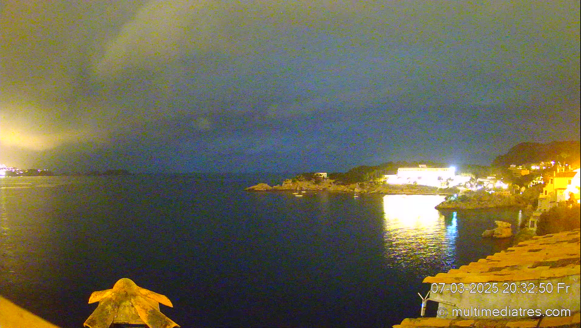 A nighttime view of a calm, dark body of water with a shoreline featuring illuminated buildings and rocky outcrops under a cloudy sky.