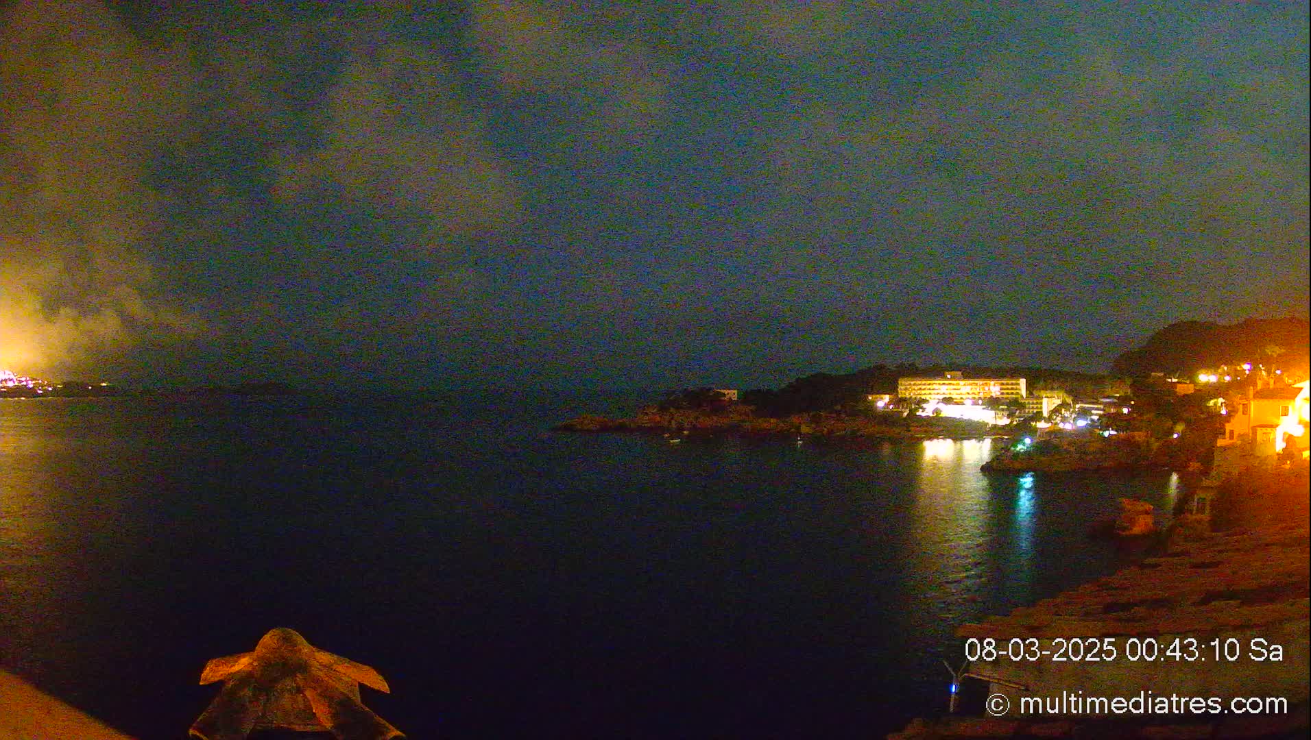 A nighttime view of a calm body of water with illuminated buildings on the opposite shore under a cloudy sky.