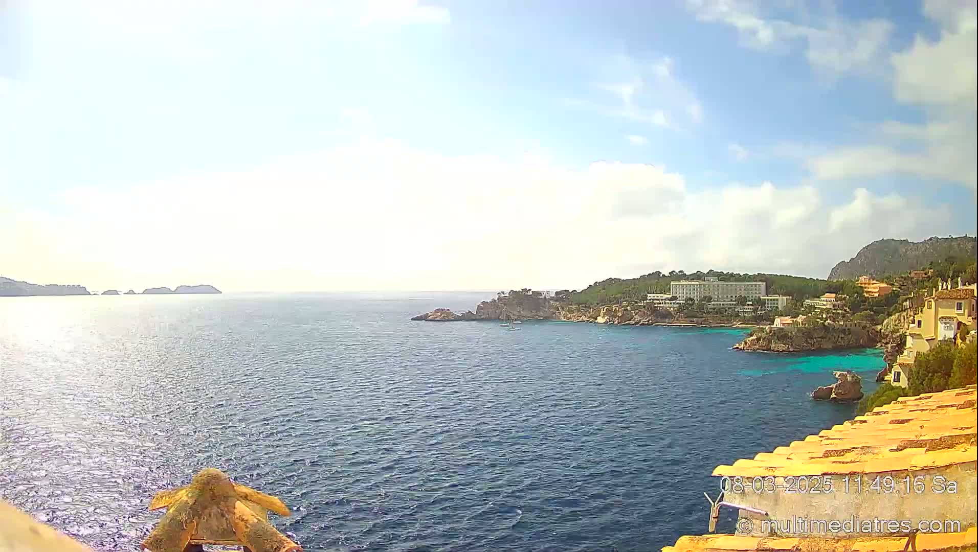 A partly sunny coastal scene shows a calm sea with a rocky shoreline featuring buildings and hotels, and small islands in the distance.