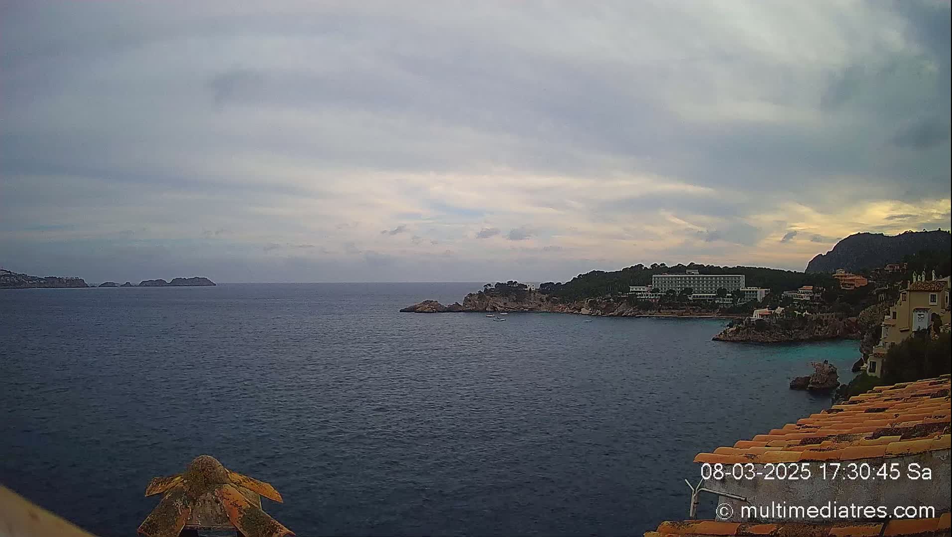 A tranquil ocean scene under a cloudy sky shows a coastline with buildings and rocky outcrops, including small islands in the distance.