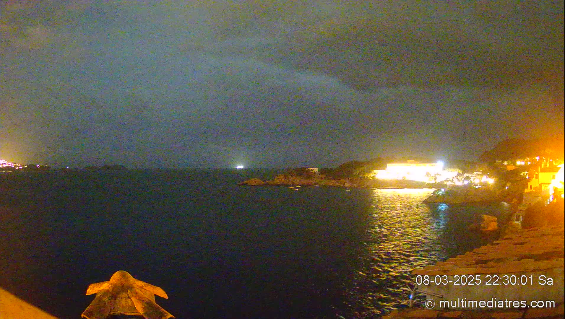 A nighttime coastal scene under a dark, cloudy sky shows a body of water reflecting the lights of buildings on the shore.