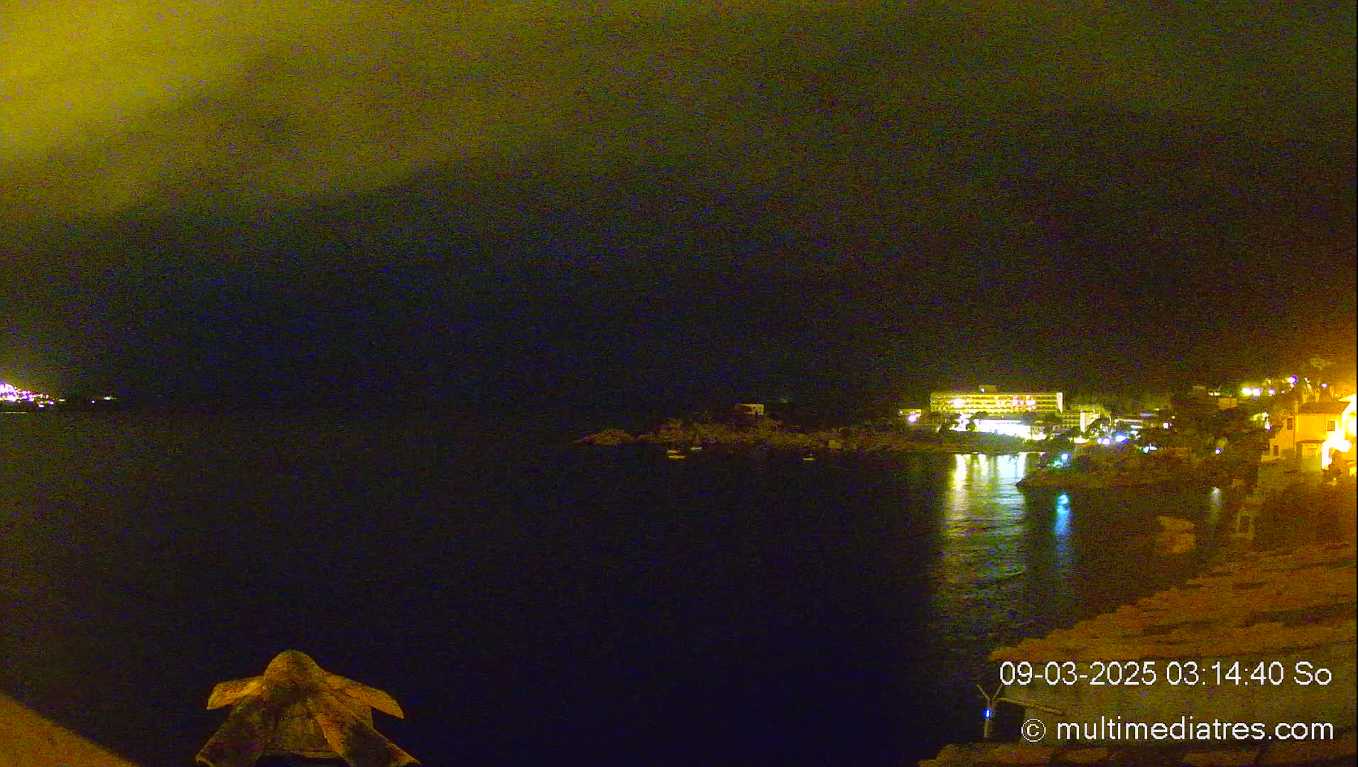 A nighttime view of a calm body of water with illuminated buildings and a landmass on the opposite shore under a dark, possibly cloudy sky.
