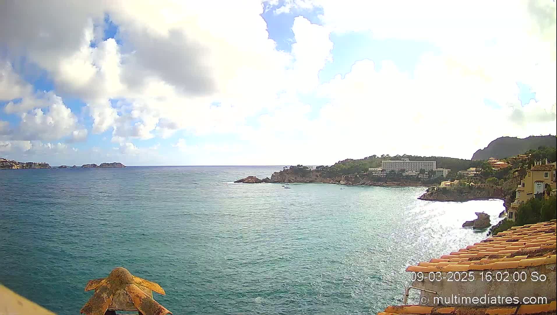 A partly cloudy day reveals a coastal scene with a calm, teal-colored sea, rocky shores, and buildings nestled along the hillside.