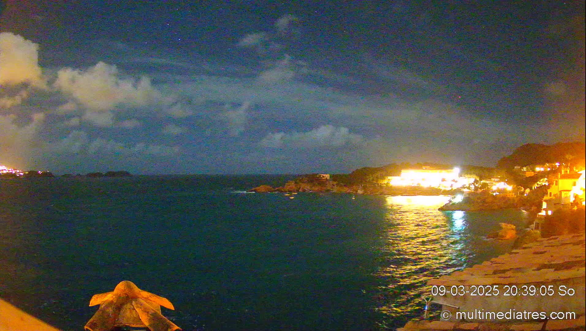 A nighttime view of a calm, dark-blue ocean with a brightly lit coastal town on the right, under a partly cloudy sky sprinkled with stars.