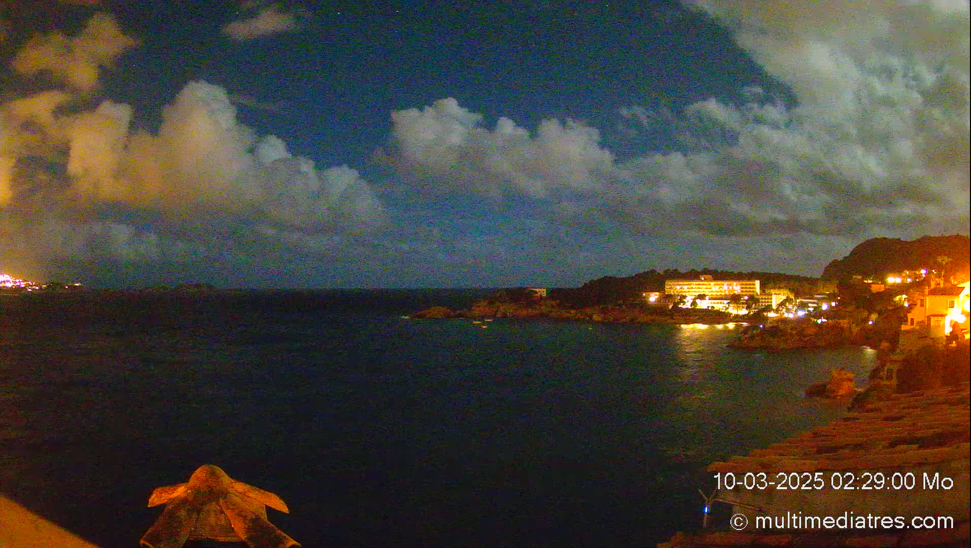 A nighttime coastal scene shows a calm, dark body of water reflecting the lights of buildings on a nearby shore under a partly cloudy sky.
