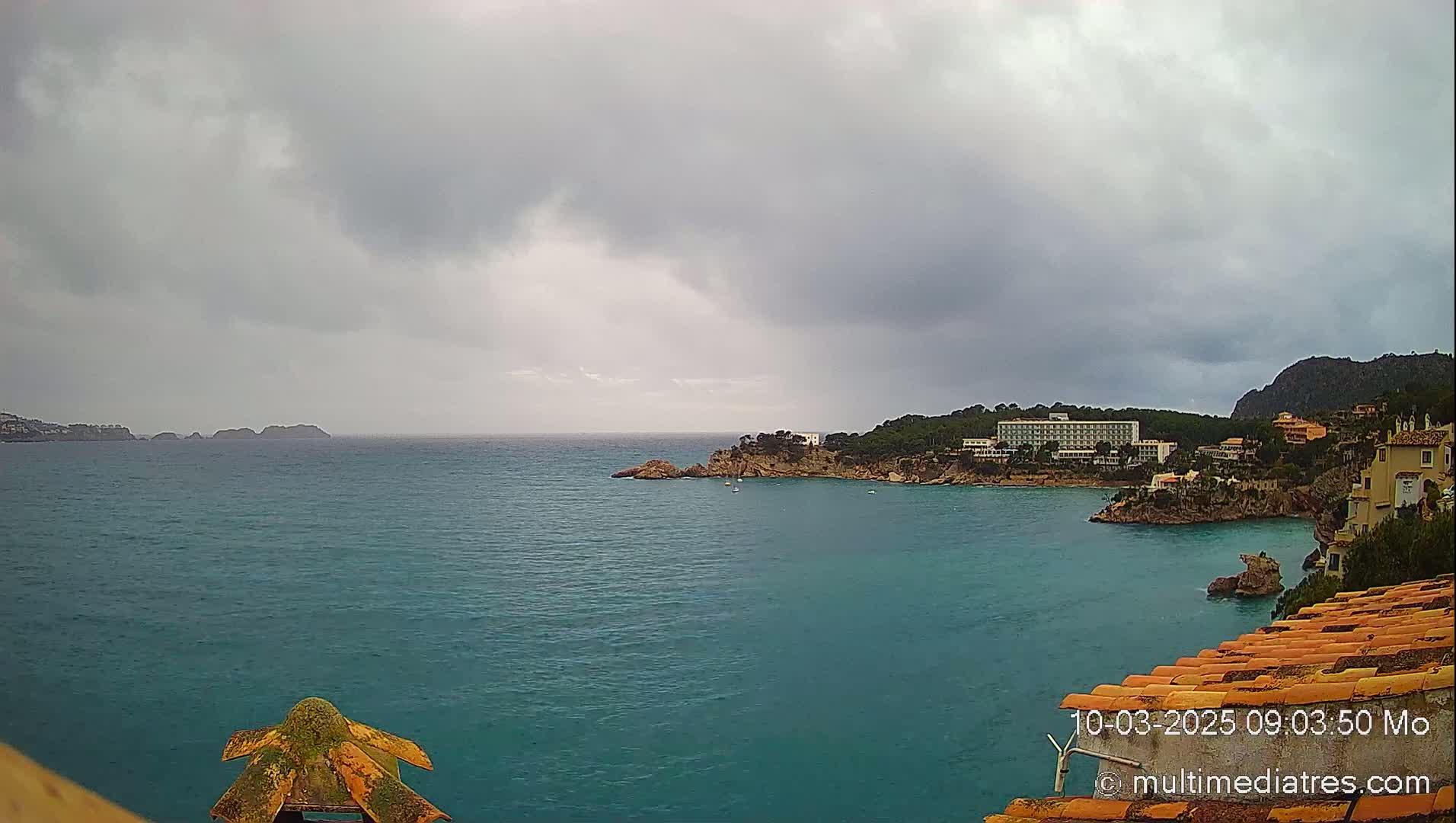 A turquoise ocean bay is seen under a cloudy, overcast sky, with buildings and trees lining the coast.