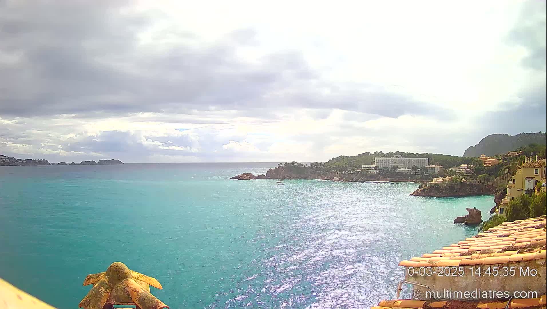 A turquoise ocean cove is partially surrounded by buildings and a rocky peninsula under a mostly cloudy sky.