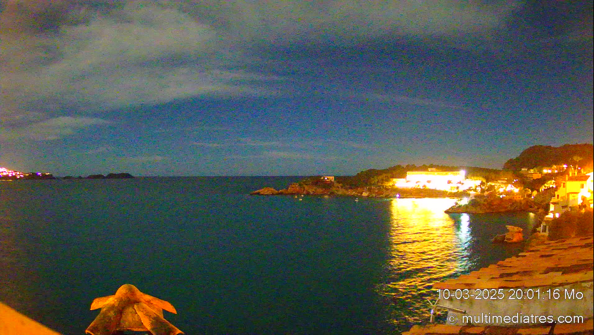 A nighttime view of a calm sea reflecting the lights of buildings on a coastal hillside under a partly cloudy sky.