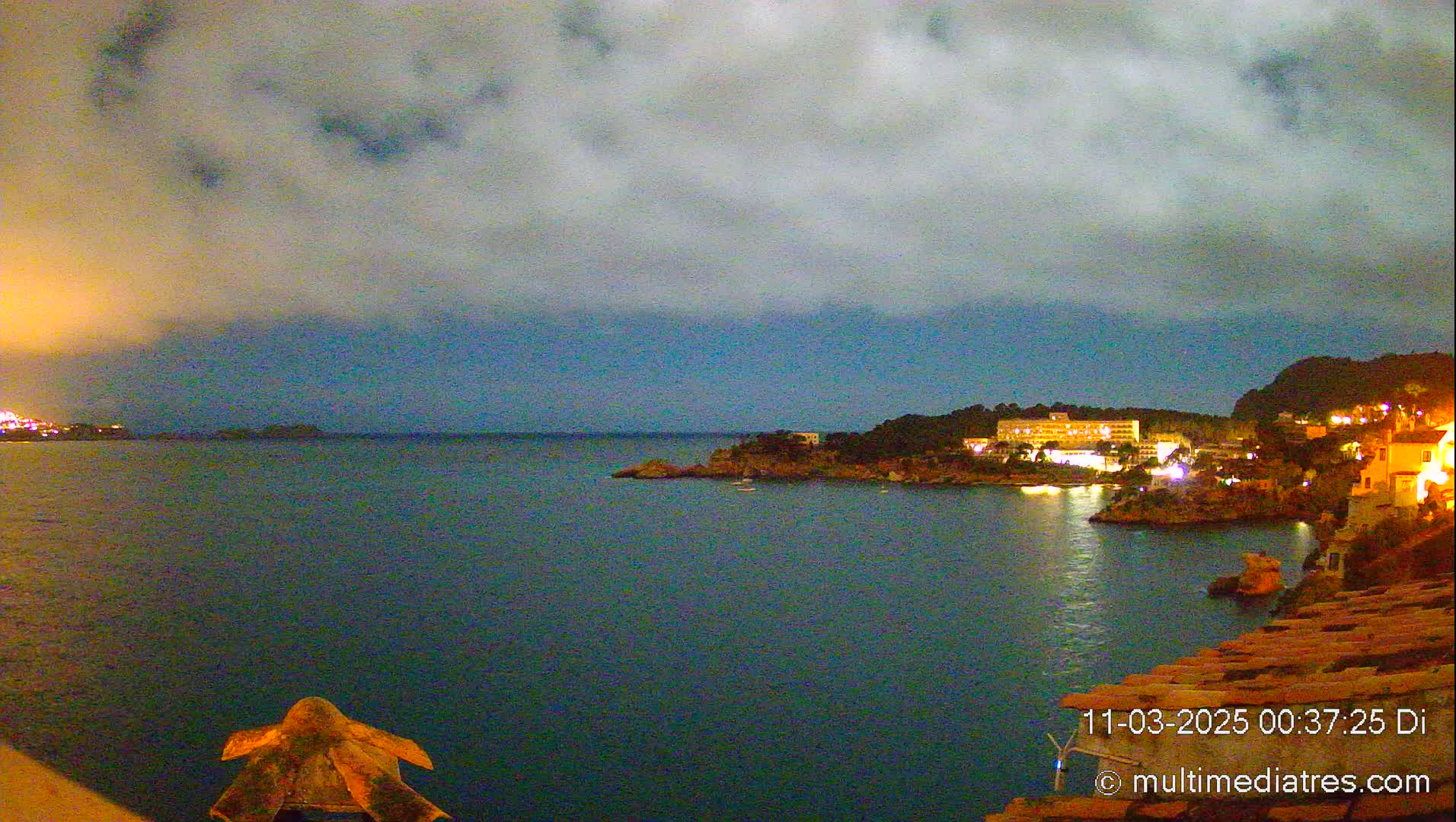 A nighttime view of a calm, dark-blue sea with a coastal town featuring lit buildings under a cloudy sky.