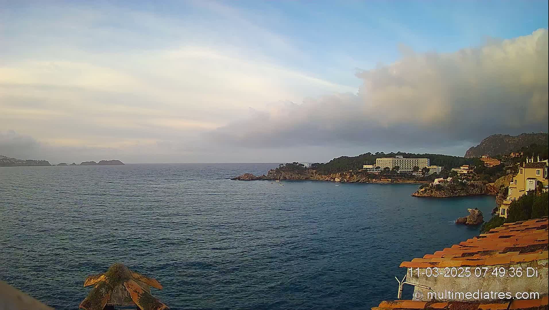 A calm, dark-blue sea laps against a rocky coastline, where buildings and a large hotel sit under a partly cloudy sky.