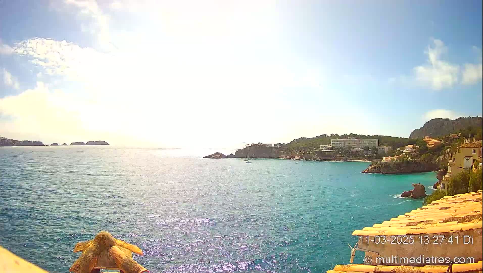 A sunny day reveals a picturesque coastal scene with a calm blue sea, rocky shores lined with buildings, and distant islands under a partly cloudy sky.