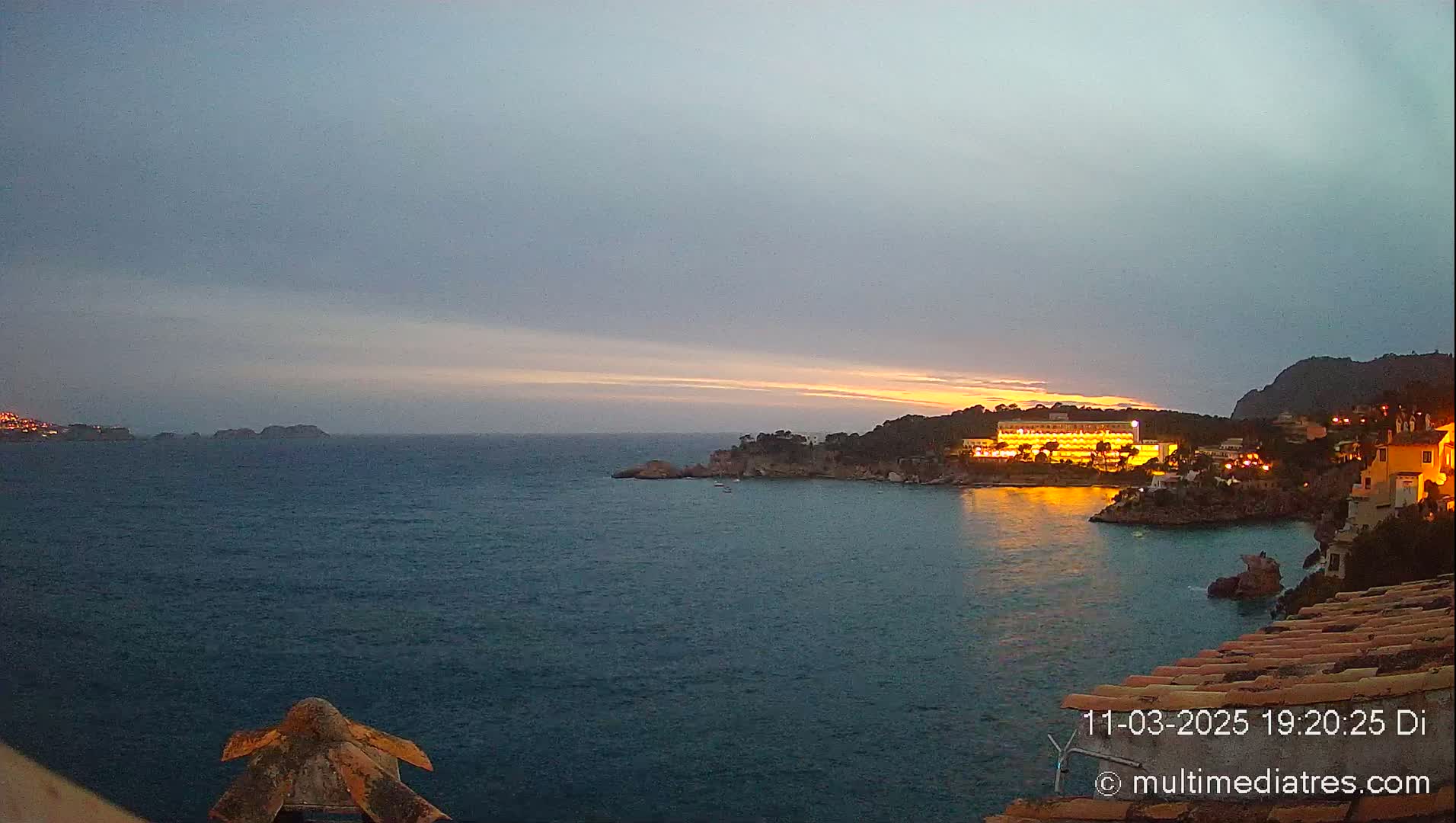 A coastal scene at dusk shows a calm sea with a brightly lit hotel and buildings on a rocky shoreline under a mostly cloudy sky.