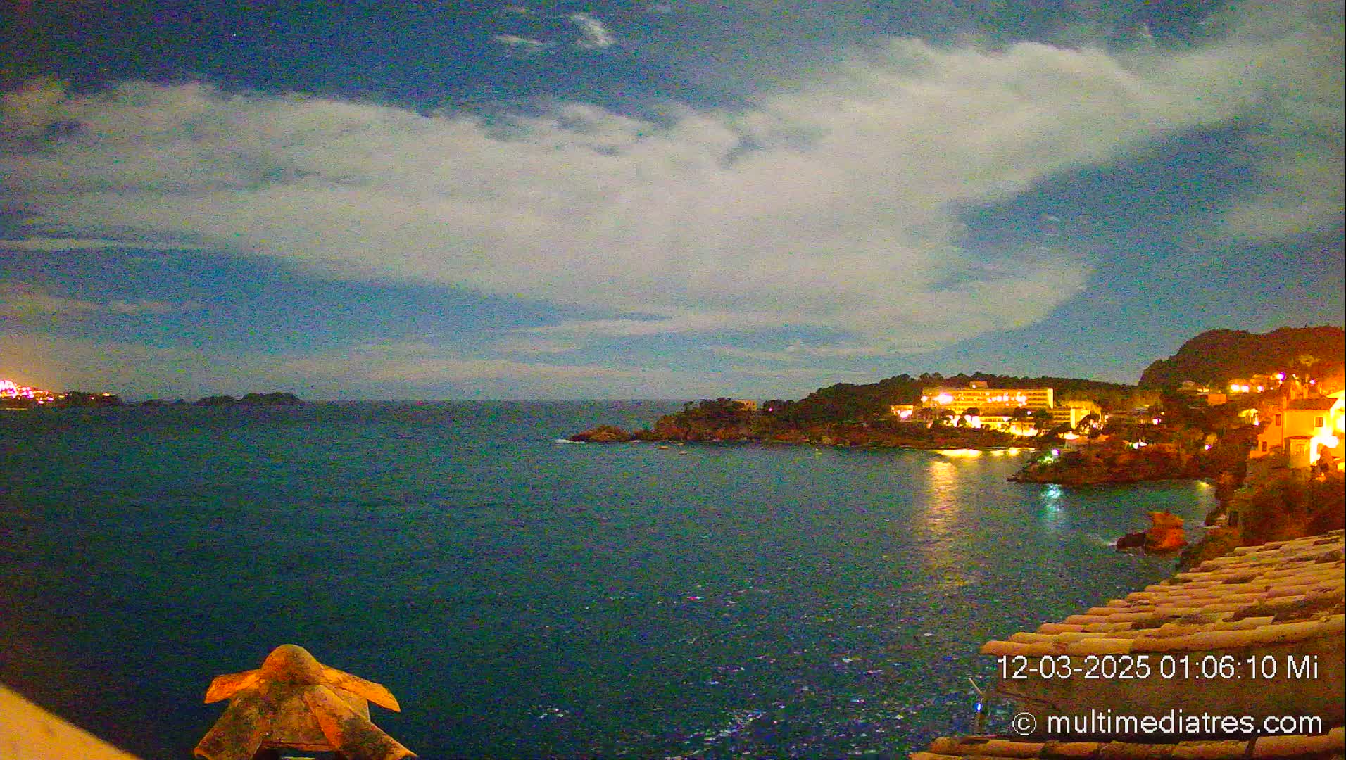A nighttime coastal scene shows a dark-blue sea under a mostly cloudy sky, with illuminated buildings and trees lining a rocky shore.