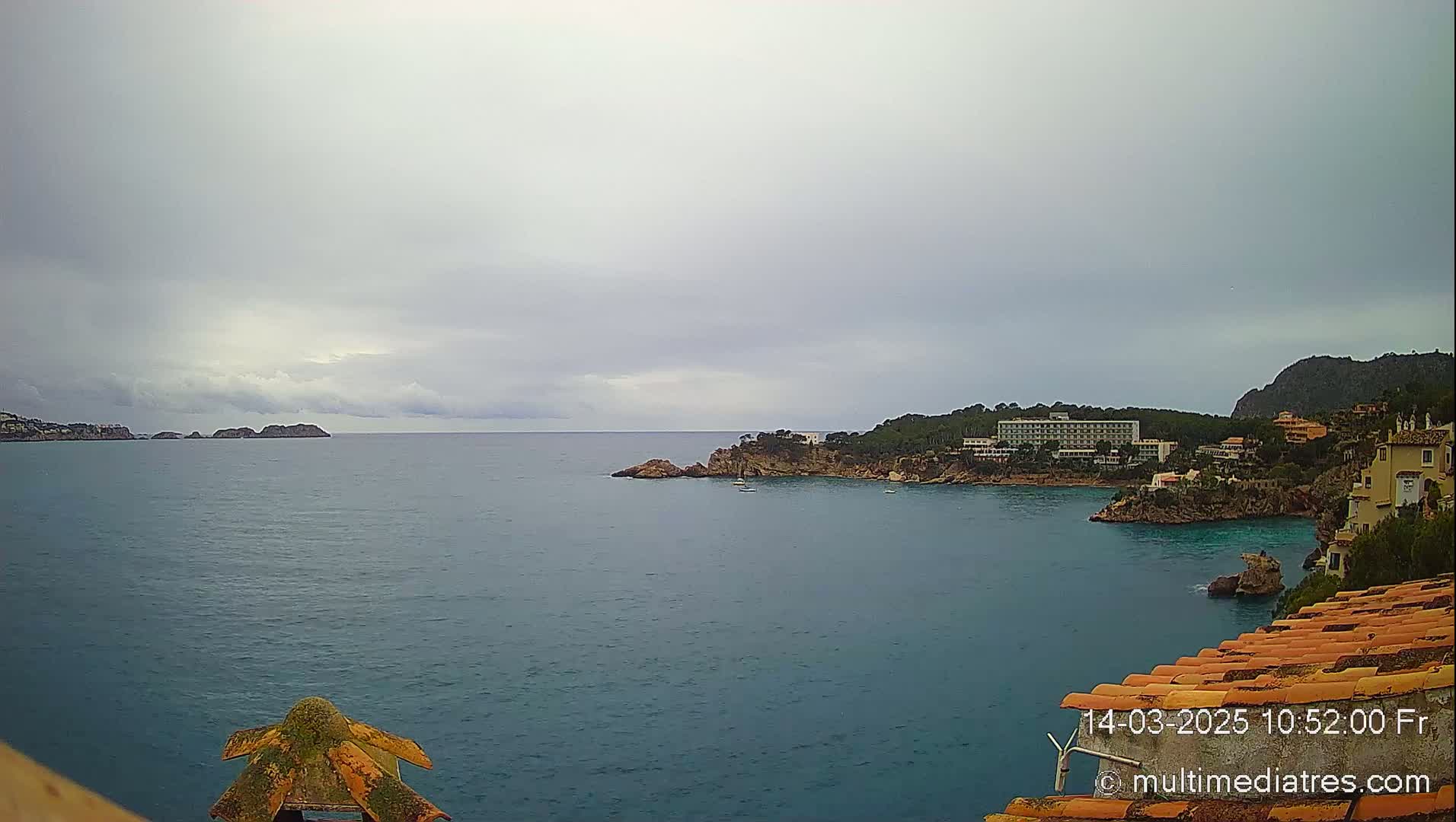 A coastal scene under an overcast sky shows a calm sea with rocky shores, some buildings, and a few small islands in the distance.