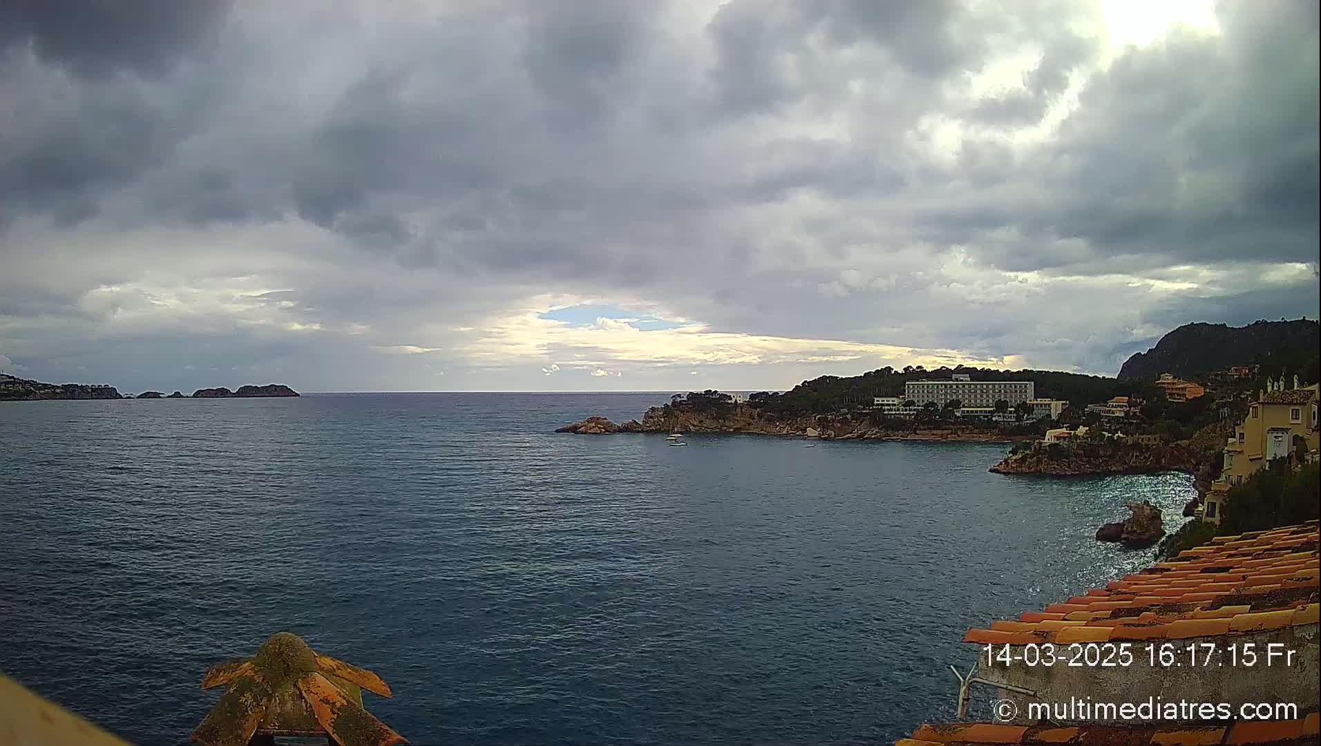 A partly cloudy day reveals a calm sea with a coastline featuring buildings, trees, and rocky outcrops.