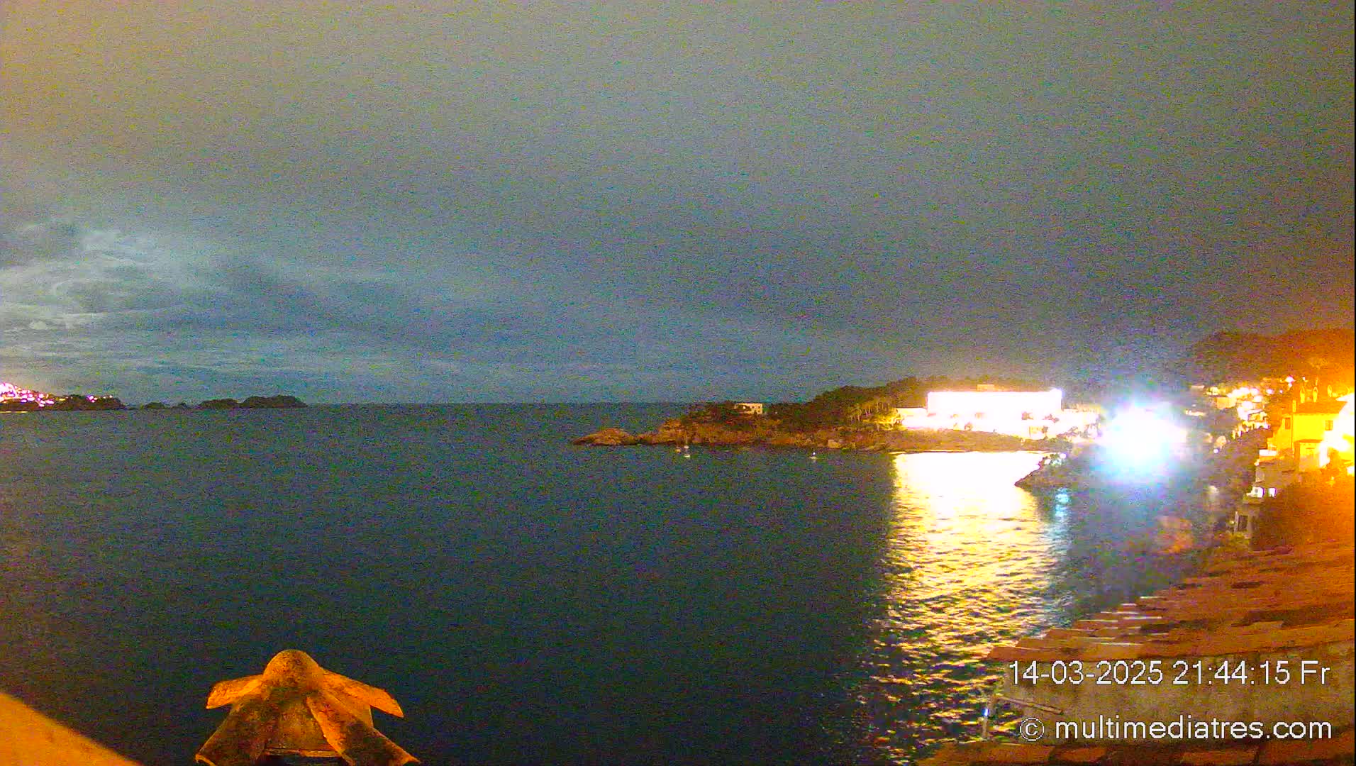 A nighttime coastal scene shows a dark body of water reflecting the lights of buildings on the shore under a cloudy sky.
