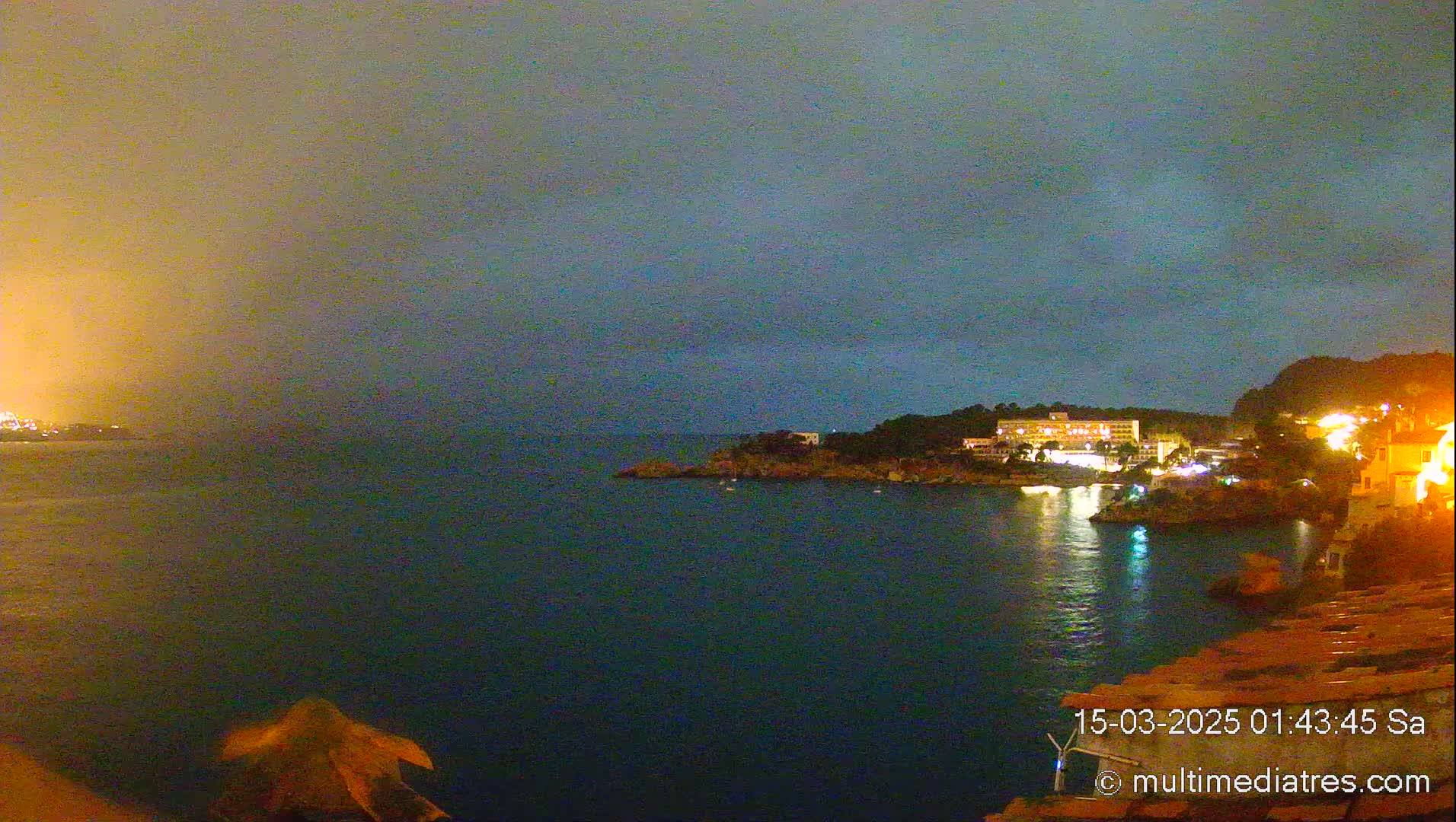 A nighttime coastal scene shows a dark body of water reflecting the lights of buildings on a nearby shore under a cloudy sky.