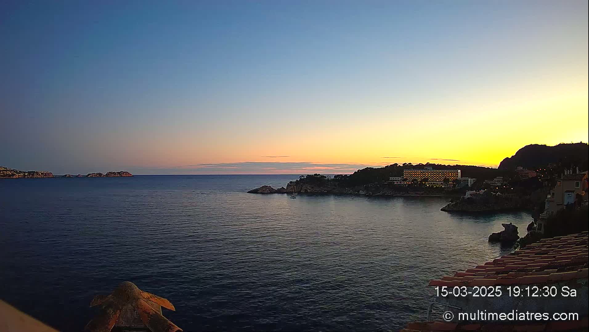 A coastal scene at sunset shows a calm sea with a hotel and buildings on a rocky shoreline, and distant islands under a clear sky.