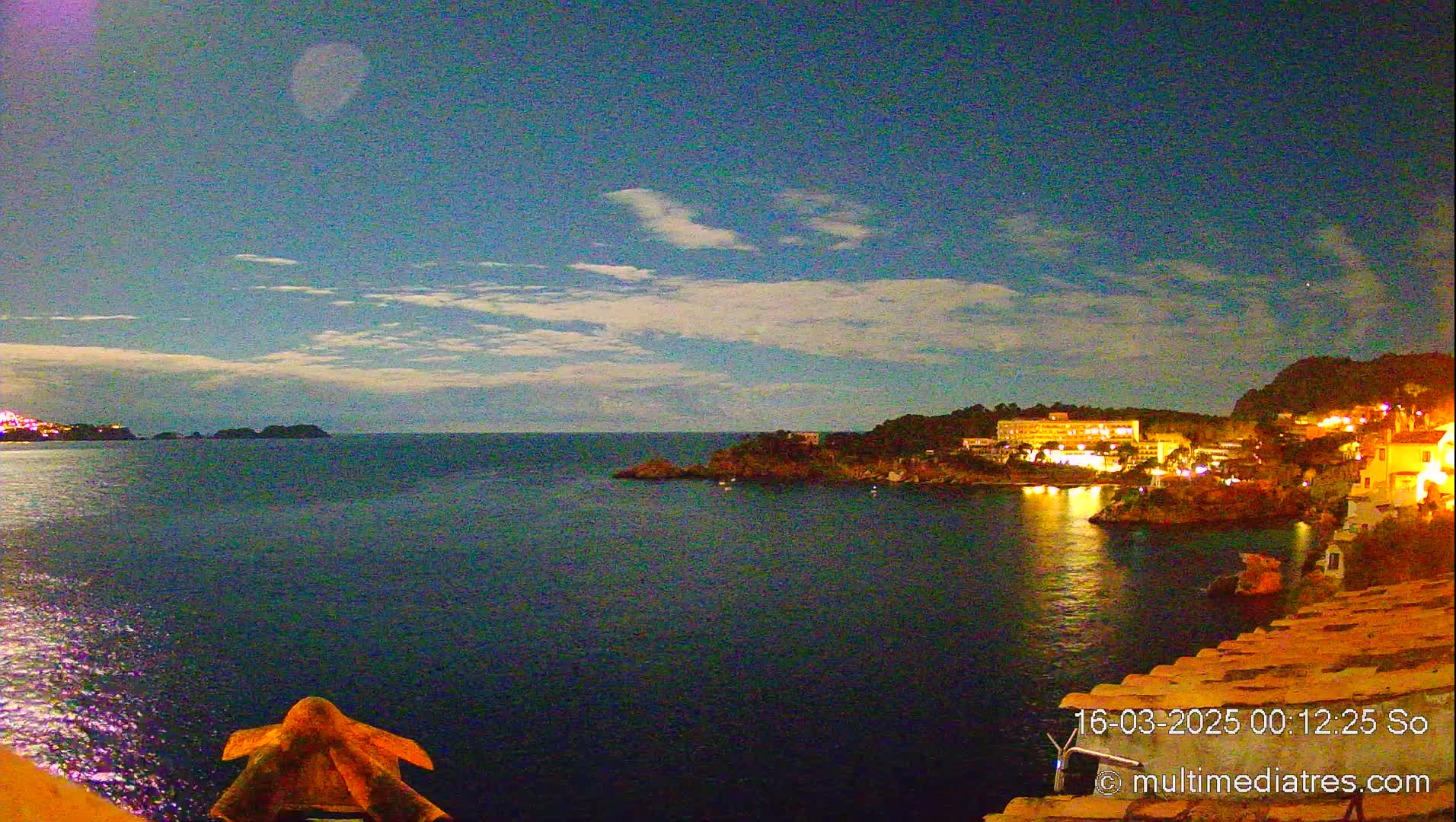 A nighttime coastal scene shows a calm, dark-blue sea reflecting the lights of buildings on a hillside under a mostly clear, twilight sky with a few clouds.