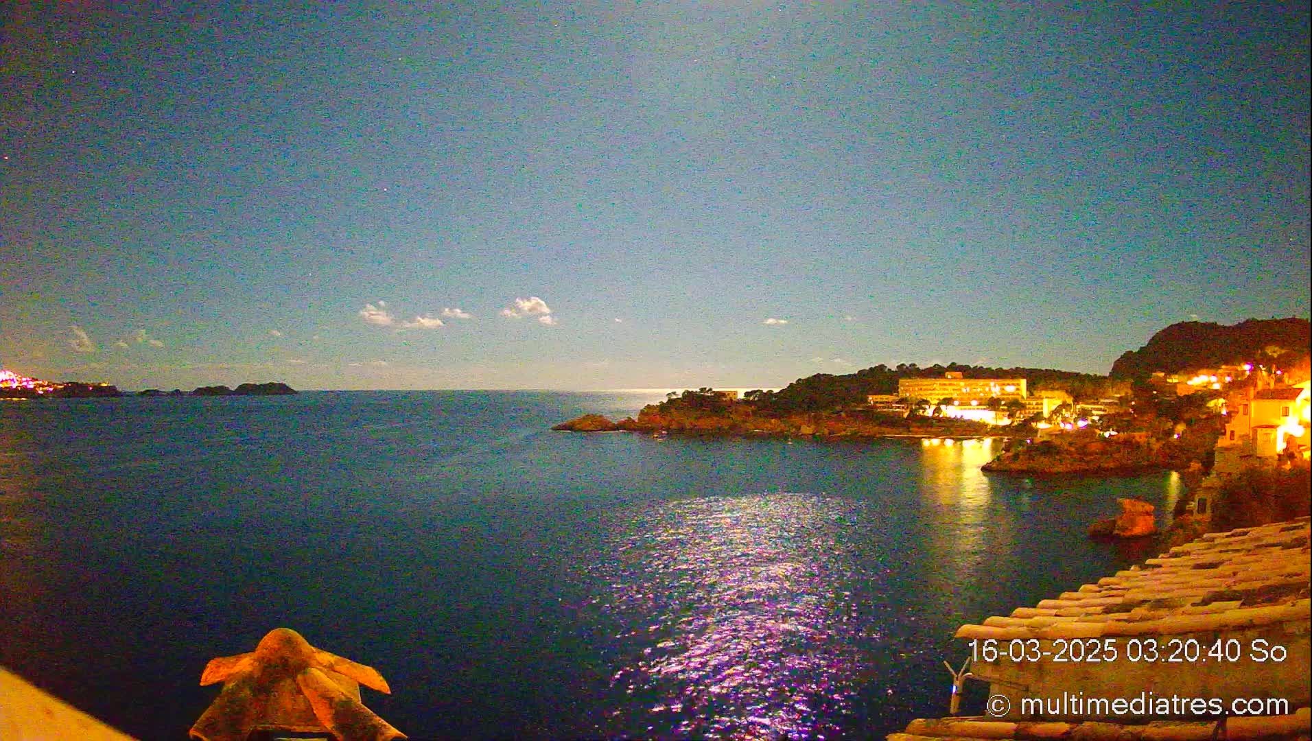 A moonlit night scene shows a calm body of water reflecting the lights of buildings on a distant shore, under a clear, dark blue sky with a few clouds.