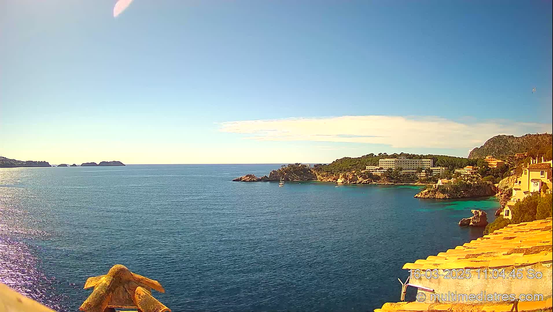 A clear, sunny day shows a coastal scene with calm blue water, rocky shorelines, and buildings nestled along a hillside.