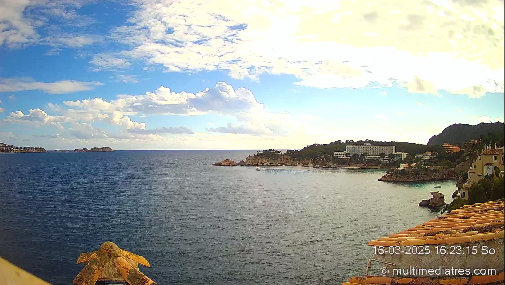 A partly cloudy day reveals a picturesque coastal scene with a calm blue sea, rocky shoreline, buildings nestled into the hillside, and small islands in the distance.