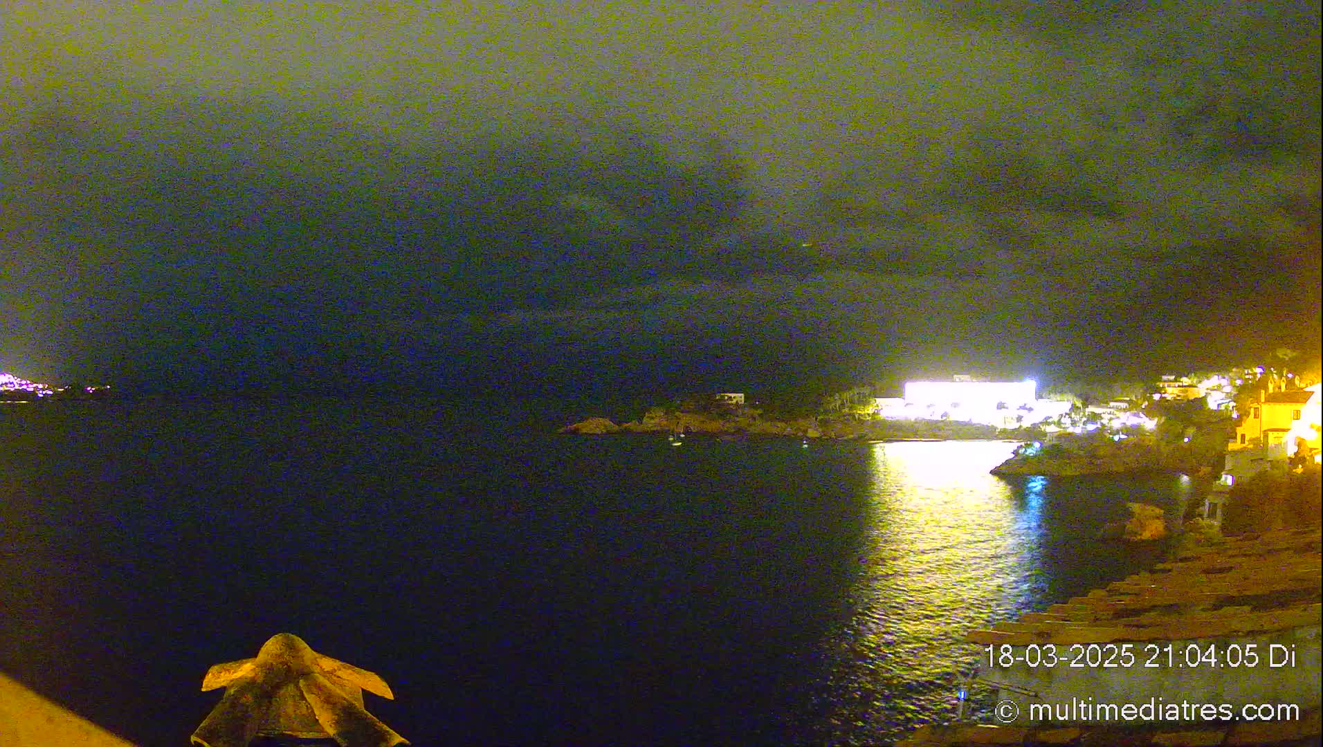 A nighttime view of a calm, dark body of water with illuminated buildings and a rocky shoreline under a cloudy sky.