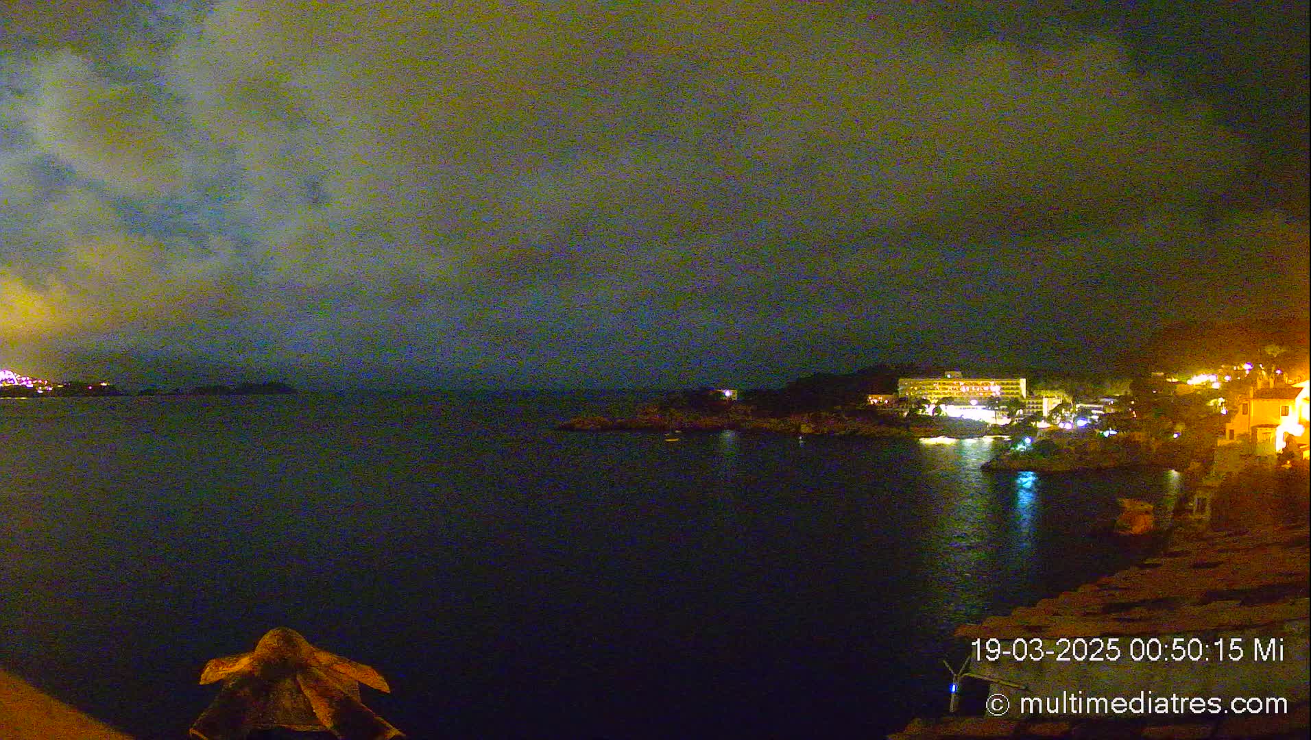 A nighttime coastal scene shows a dark body of water with illuminated buildings and a cloudy sky.