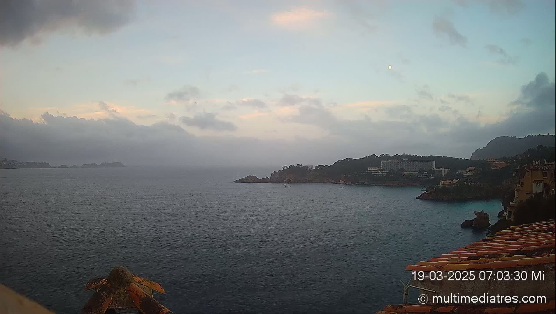 A hazy sunrise over a calm bay with a small town and distant islands visible under a mostly cloudy sky.