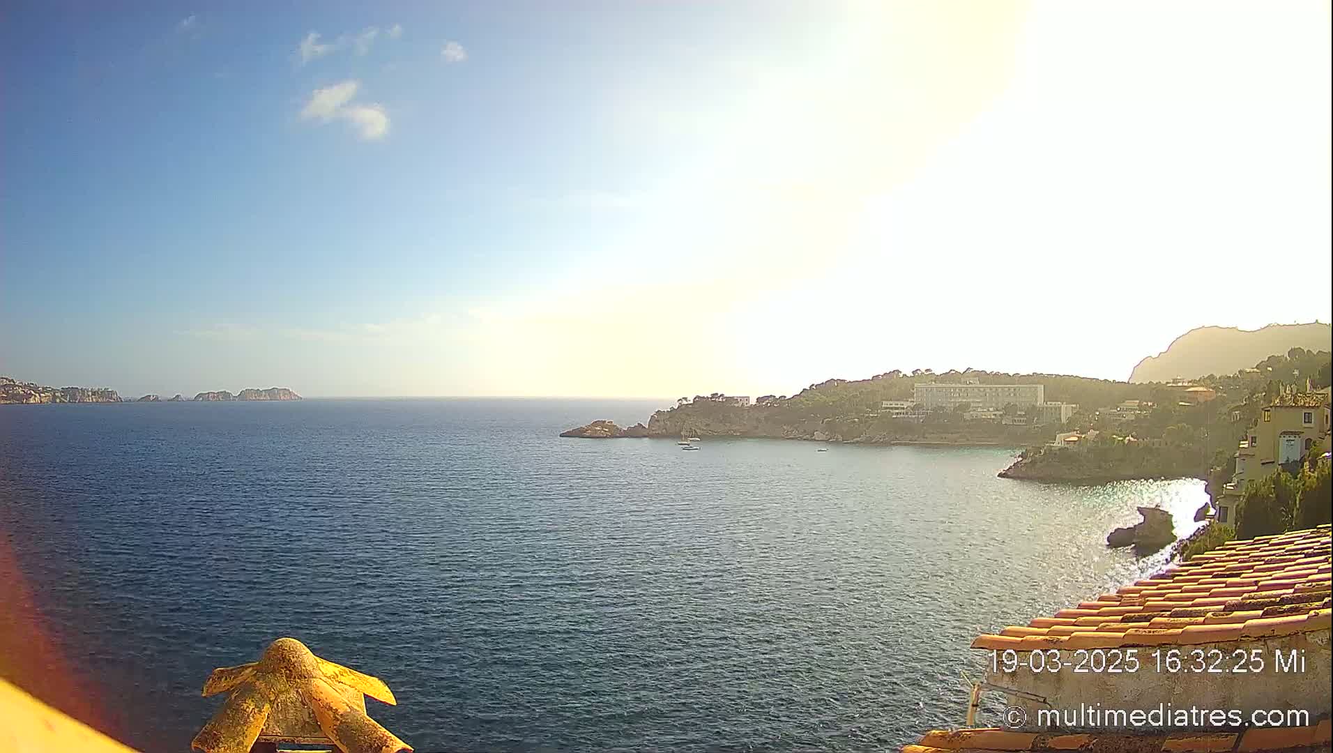 A sunny coastal scene shows a calm, blue sea with rocky shores and buildings on a hillside, under a mostly clear sky.