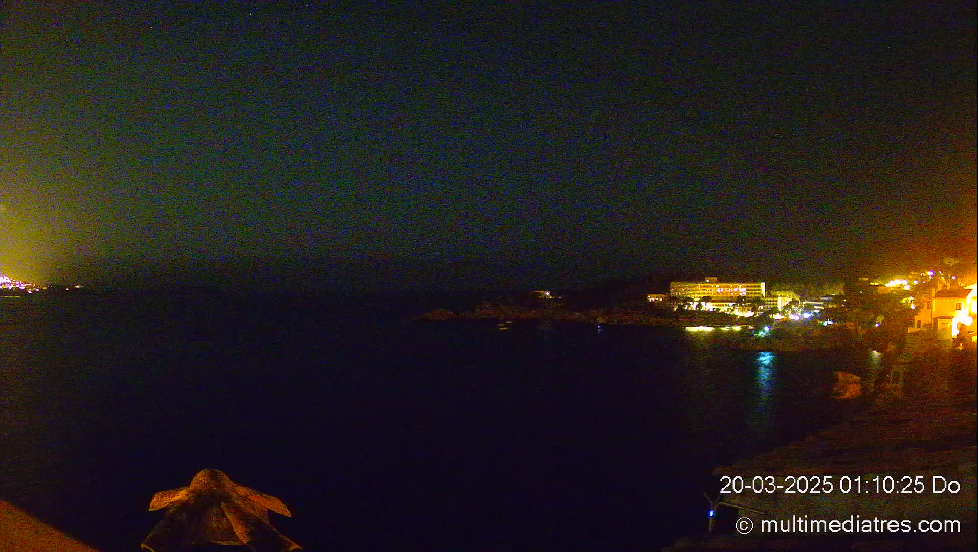 A nighttime view of a dark body of water with illuminated buildings and a rocky shore under a clear, starry sky.