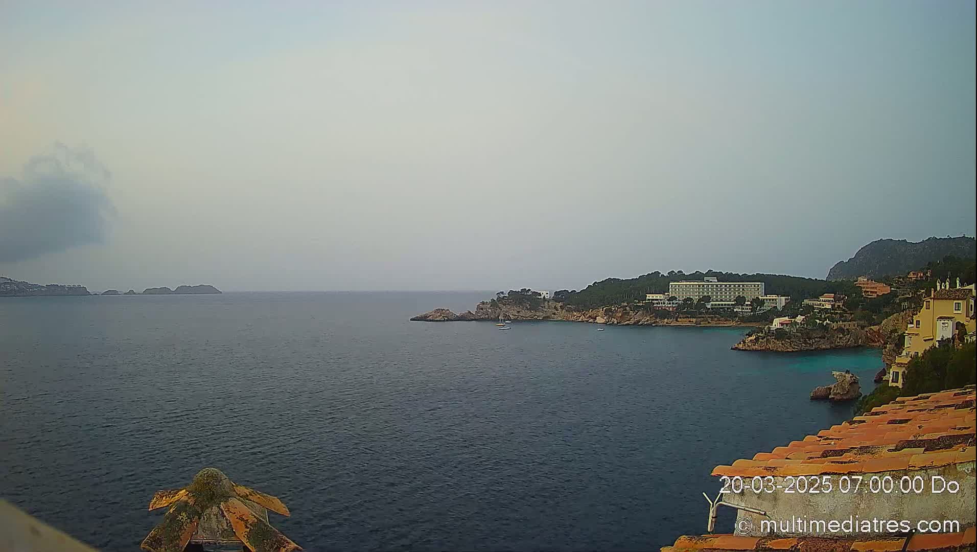 A hazy, overcast day reveals a coastal scene with a calm sea, rocky inlets, a large hotel, and several buildings nestled along the shoreline.