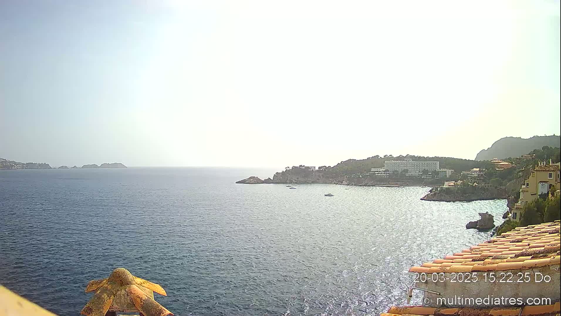 A hazy, sunny day overlooks a calm sea with a coastline featuring buildings and rocky outcrops.